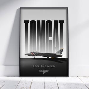 May include: A framed poster featuring a Tomcat fighter jet. The word "TOMCAT" is printed in large, bold, black and white letters above the aircraft. Below the jet, the text "FEEL THE NEED" and the Grumman logo are displayed.