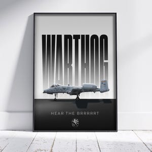 May include: Framed print featuring a gray A-10 Thunderbolt II aircraft, with the word "WARTHOG" in large, bold, black and white letters above it. Below the aircraft, the text reads "HEAR THE BRRRRRT". The print is in a black frame.