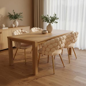 Könnte beinhalten: Ein Esstisch aus hellem Holz mit vier cremefarbenen Stühlen. Die Stühle haben goldene Beine und ein einzigartiges, blasenartiges Rückendesign. Eine Vase mit Grün steht auf dem Tisch, und ein Holzschrank ist im Hintergrund zu sehen.