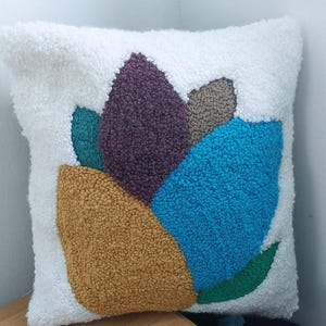 May include: A square throw pillow featuring a white textured, looped pile background with a central abstract design. The pattern consists of large, organic shapes in solid colors including mustard yellow, deep purple, bright blue, teal, and gray.