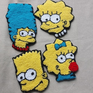 以下が含まれることがあります： 手作りのカラフルな刺繍キャラクターパッチ4枚。アニメテレビ番組の、マージ、リサ、バート、マギー・シンプソンの顔が描かれています。各パッチは黒いアウトラインで、ニュートラルな背景に配置されています。