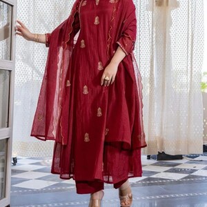 May include: A burgundy three-piece outfit, comprising a long tunic, trousers, and a sheer scarf. The tunic and scarf feature gold embroidery. The outfit is worn with clear strap heels.