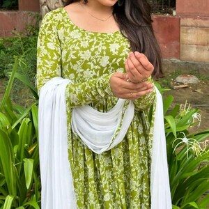 Puede incluir: Un vestido verde de manga larga con un estampado floral blanco y una bufanda blanca a juego. El vestido tiene un cuello redondo y est&aacute; hecho de una tela ligera. La bufanda est&aacute; drapeada sobre los hombros.