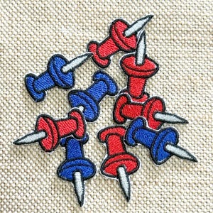 May include: A collection of embroidered patches shaped like push pins in vibrant red and blue colors. Each patch features a circular head and a sharp, silver-toned needle point, rendered with detailed stitching and black outlines throughout.