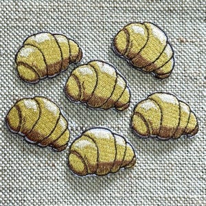 Peut inclure: Six écussons brodés en forme de croissants dorés. Chaque pièce présente des coutures détaillées dans des tons de jaune et de brun pour imiter la texture feuilletée de la pâtisserie, avec une forme en croissant incurvée et des contours sombres.