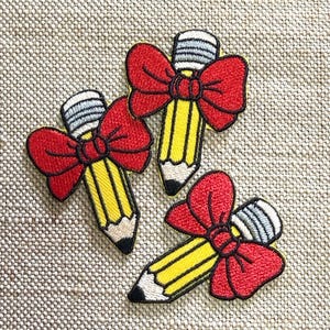 Puede incluir: Tres parches bordados, cada uno con un lápiz amarillo con punta negra y goma de borrar gris, acentuados por un gran lazo rojo brillante atado alrededor del centro del lápiz. Los parches están dispuestos en grupo sobre una superficie de tela.