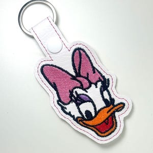 May include: A white keychain featuring an embroidered Daisy Duck design. The keychain has a pink bow, orange beak, and black and purple details. It includes a silver metal ring.