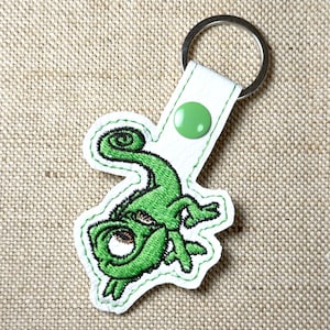 May include: A green embroidered chameleon keychain with a white leather strap and a silver key ring. The chameleon has a detailed design with black outlines and a green button closure. The keychain is on a tan fabric background.