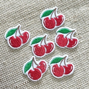 Può includere: Sei toppe ricamate a forma di ciliegia, raffiguranti due ciliegie rosso brillante con una foglia verde e gambi marroni. Ogni toppa ha un bordo bianco ed è composta da una coppia di ciliegie unite dai gambi, con dettagliate cuciture rosse.