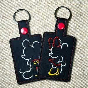 May include: Two black keychains featuring Mickey and Minnie Mouse designs. The keychains have a rectangular shape with a red button and stitching. Mickey is outlined in white with yellow accents, and Minnie is outlined in red and white with yellow accents.