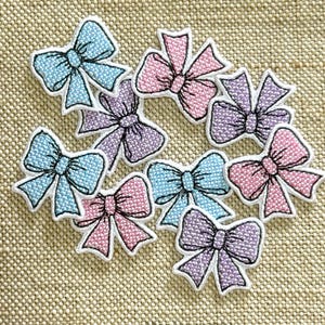 Puede incluir: Una colección de nueve parches bordados con forma de lazo en tonos pastel azul claro, rosa suave y lavanda. Cada lazo presenta un borde blanco y detalles cosidos en negro para definir los pliegues de la tela y el nudo central.
