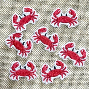 May include: Seven small embroidered crab patches featuring bright red bodies with white borders and black eyes. Each patch depicts a crab with raised claws and multiple legs, arranged in a scattered pattern on a textured beige fabric surface.