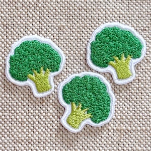 Puede incluir: Tres parches bordados con forma de ramilletes de brócoli. Cada parche presenta una parte superior de color verde vibrante con una superficie texturizada y un tallo de color verde amarillento claro, bordeado por un contorno blanco.