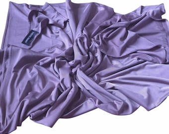 May include: A light purple fabric with a smooth, slightly reflective surface and a soft, draped texture. A black rectangular tag is attached to the fabric, featuring the white text "LIQUID JERSEY" printed in a clear, sans-serif font.