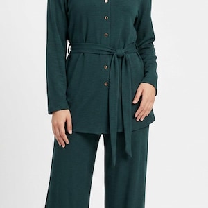 Puede incluir: Un conjunto de dos piezas de color verde oscuro que consta de una túnica de manga larga con botones, un cinturón de lazo a juego y pantalones anchos. La túnica presenta una hilera de pequeños botones metálicos en la parte delantera y un pañuelo.
