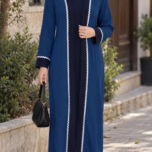 May include: A long-sleeved navy blue open-front duster coat featuring white decorative trim along the edges and cuffs, worn over a matching navy blue dress and a navy blue hijab, paired with white sneakers and a small black handbag.