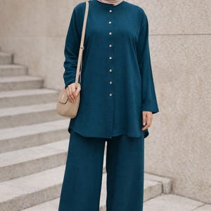 May include: A teal long-sleeved button-down tunic and matching wide-leg trousers set. The outfit is paired with a beige hijab, a beige crossbody bag, and beige open-toe sandals. The text "CRESCENT COVERS" is visible in the top right corner.