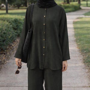 May include: A dark olive green long-sleeved button-down shirt and matching wide-leg trousers. The shirt features a round neckline and a black hijab. A black woven shoulder bag and brown sunglasses are held. The text CRESCENT COVERS is visible.