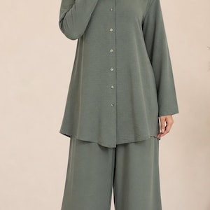May include: A sage green two-piece set consisting of a long-sleeved button-down tunic with a round neckline and matching wide-leg trousers. The tunic features a full-length front button placket with small buttons and a curved hemline.