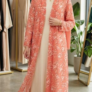 以下が含まれることがあります： クリーム色のドレスの上に、長い、流れるようなピーチ色のフローラルプリントのカーディガン。カーディガンは長袖で、繊細な白い花柄です。お揃いのピーチ色のヒジャーブを着用しています。