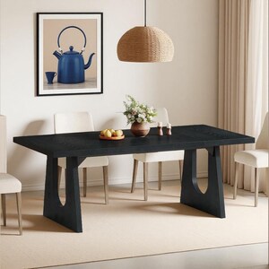 May include: A black rectangular dining table with a unique leg design, complemented by cream-colored chairs. A framed picture of a blue teapot hangs on the wall. A woven pendant light hangs above the table, and a vase of flowers sits on the table.