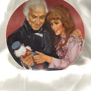 May include: A vintage porcelain plate featuring a painting of an older man and a young girl holding a nutcracker doll. The plate has a red background and the figures are dressed in period clothing.