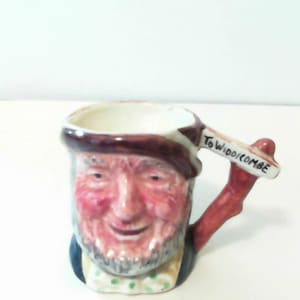May include: A small ceramic mug with a cartoon face and a handle shaped like a branch. The mug has the words "To Widdicombe" written on it.