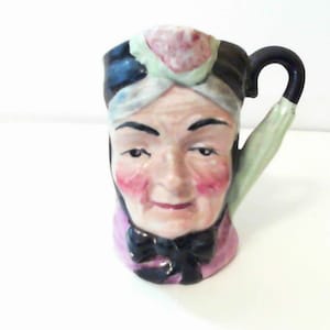 May include: A ceramic pitcher shaped like a person's head with a black turban and a green and white patterned scarf. The pitcher has a handle and a light pink and green base.