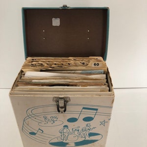 Könnte beinhalten: Vintage weiße Schallplatten-Aufbewahrungsbox mit türkisblauem Deckel und Illustration von Noten und Band. Die Box ist geöffnet und zeigt zahlreiche Schallplattenhüllen. Die Box hat einen Metallverschluss und ist leicht abgenutzt.