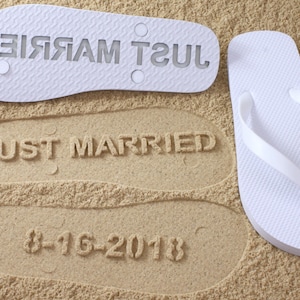 May include: White flip flops with the words "Just Married" printed on the soles. The flip flops are on a sandy beach with the words "Just Married" and the date "8-16-2018" imprinted in the sand.