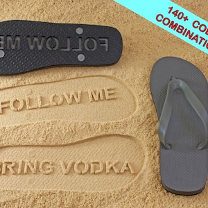 May include: Two black flip flops with the words "FOLLOW ME" printed on the soles. One flip flop is laying on the sand, the other is standing upright. The flip flops are on a sandy beach.