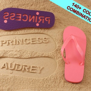 May include: Purple flip flop with the word "Princess" printed in pink on the sole. The flip flop is laying on sand with the words "Princess" and "Audrey" imprinted in the sand.