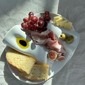 May include: A white, multi-sectioned serving platter holds a selection of food items. The platter contains red grapes, sliced cheese, olives, sliced meat, figs, bread, and a small pool of oil.