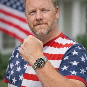 Op de afbeelding: Een polshorloge met een zwarte wijzerplaat en zilveren wijzers wordt getoond. Het horloge heeft een roestvrijstalen band. De persoon draagt een rood, wit en blauw T-shirt met een Amerikaans vlagontwerp.