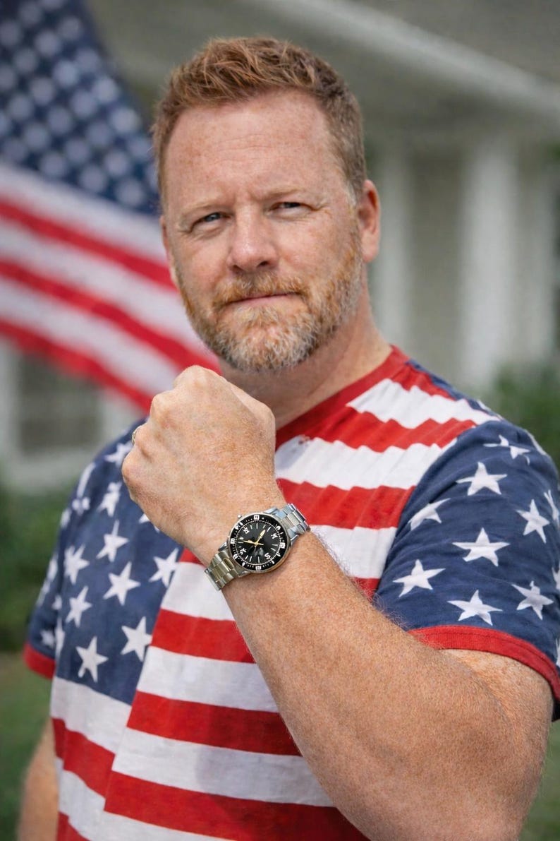Op de afbeelding: Een polshorloge met een zwarte wijzerplaat en zilveren wijzers wordt getoond. Het horloge heeft een roestvrijstalen band. De persoon draagt een rood, wit en blauw T-shirt met een Amerikaans vlagontwerp.