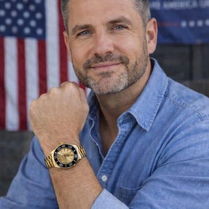Pu&ograve; includere: Un orologio da polso dorato con lunetta nera e cinturino dorato. Il quadrante dell'orologio ha uno sfondo chiaro con numeri e indicatori neri. La persona indossa una camicia di jeans blu.