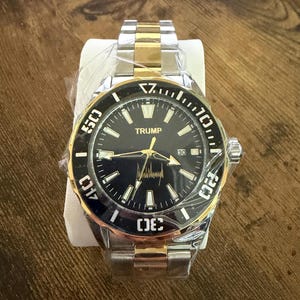 Op de afbeelding: Een tweekleurig polshorloge met een zwarte wijzerplaat en gouden accenten. Het horloge heeft het woord "TRUMP" op de wijzerplaat, samen met een handtekening. Het heeft een roestvrijstalen band en een draaibare ring met witte cijfers.
