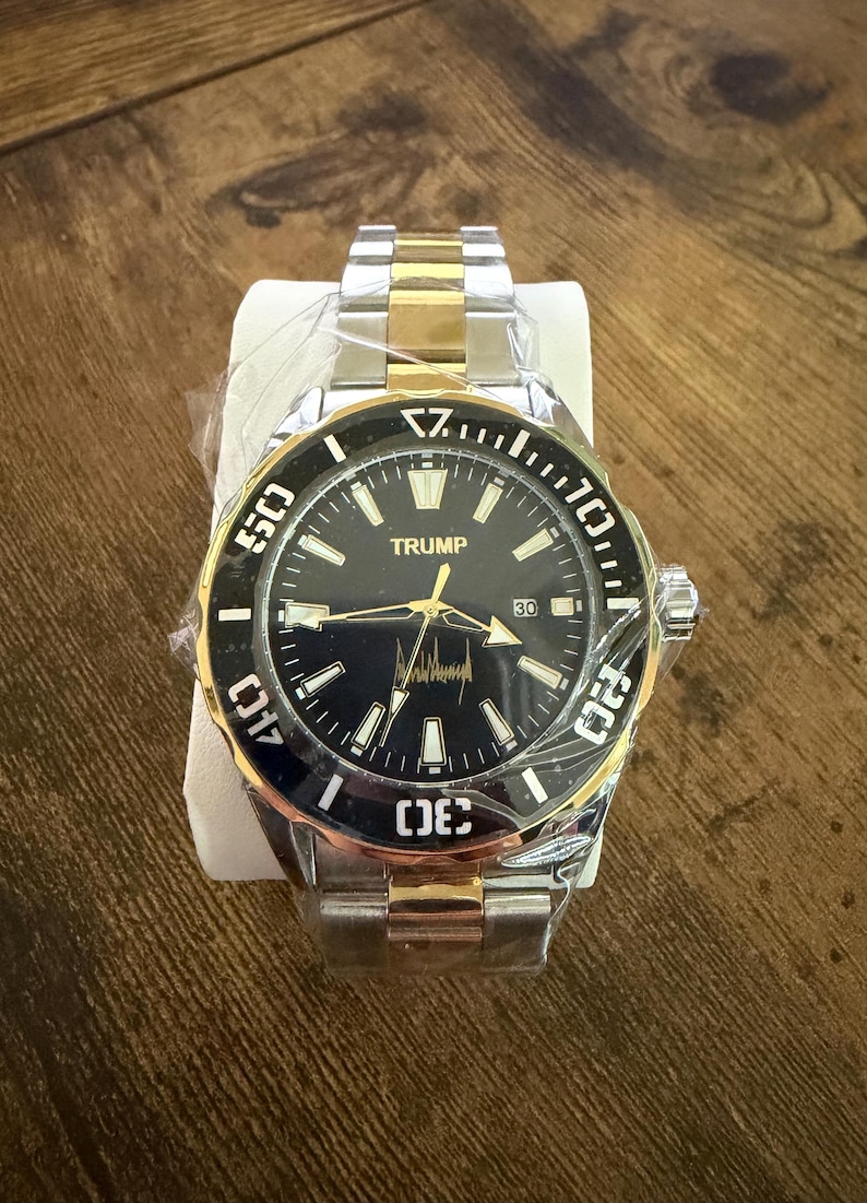 Op de afbeelding: Een tweekleurig polshorloge met een zwarte wijzerplaat en gouden accenten. Het horloge heeft het woord "TRUMP" op de wijzerplaat, samen met een handtekening. Het heeft een roestvrijstalen band en een draaibare ring met witte cijfers.