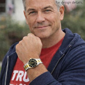 Op de afbeelding: Een goud-zilveren polshorloge met een zwarte wijzerplaat wordt om een pols getoond. Het horloge heeft een gouden bezel en een zilveren en gouden band. De persoon draagt een rood shirt met het woord "TRU" en de cijfers "= *202" erop.