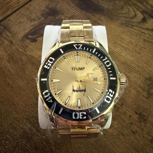Pu&ograve; includere: Orologio da polso color oro con lunetta nera e quadrante dorato. L'orologio presenta il nome "TRUMP" sul quadrante, insieme a una firma. L'orologio &egrave; nuovo e ancora avvolto nella plastica. Anche il cinturino &egrave; color oro.