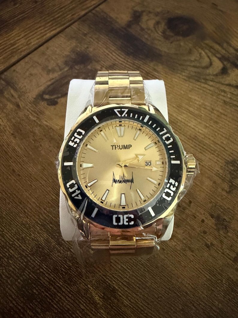 Pu&ograve; includere: Orologio da polso color oro con lunetta nera e quadrante dorato. L'orologio presenta il nome "TRUMP" sul quadrante, insieme a una firma. L'orologio &egrave; nuovo e ancora avvolto nella plastica. Anche il cinturino &egrave; color oro.