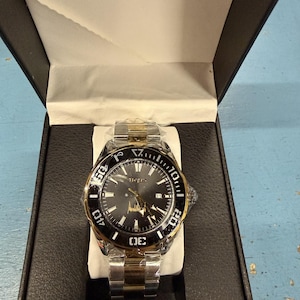 Op de afbeelding: Een tweekleurig polshorloge met een zwarte wijzerplaat en een goudkleurige lunette, gepresenteerd in een zwarte doos. Het horloge heeft een datumweergave en een roestvrijstalen band met gouden accenten. Het horloge is nieuw en ongedragen.