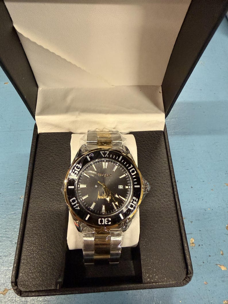 Op de afbeelding: Een tweekleurig polshorloge met een zwarte wijzerplaat en een goudkleurige lunette, gepresenteerd in een zwarte doos. Het horloge heeft een datumweergave en een roestvrijstalen band met gouden accenten. Het horloge is nieuw en ongedragen.