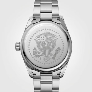 Op de afbeelding: Roestvrijstalen polshorloge met zilveren band. De wijzerplaat van het horloge heeft een gedetailleerde gravure van de Amerikaanse adelaar en de woorden "E Pluribus Unum". Het horloge heeft een ronde wijzerplaat en een zilveren kroon.