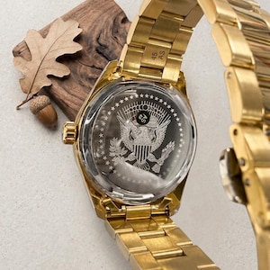 Pu&ograve; includere: Un orologio da polso dorato con cinturino in metallo. Il quadrante dell'orologio presenta un'incisione dettagliata di un emblema di aquila e scudo. L'orologio &egrave; esposto con un elemento in legno e una foglia.