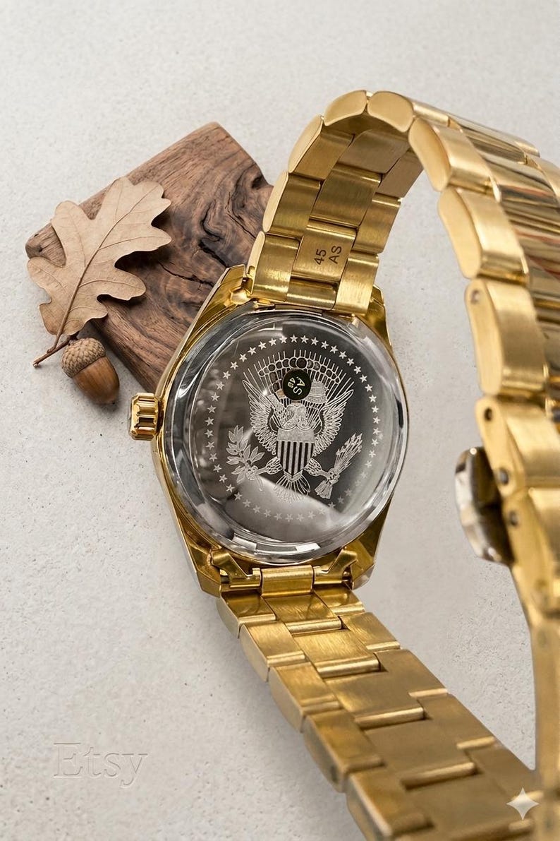 Pu&ograve; includere: Un orologio da polso dorato con cinturino in metallo. Il quadrante dell'orologio presenta un'incisione dettagliata di un emblema di aquila e scudo. L'orologio &egrave; esposto con un elemento in legno e una foglia.