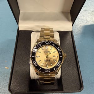 Pu&ograve; includere: Orologio da polso color oro con lunetta nera, presentato in una scatola nera. Il quadrante dell'orologio &egrave; dorato con la scritta "TRUMP" in alto. L'orologio ha un cinturino dorato ed &egrave; nuovo nella sua confezione.