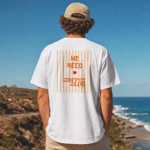 Op de afbeelding: Een wit T-shirt met korte mouwen en een rugprint van verticale lichtoranje strepen. De tekst WE NEED COFFEE CLUB staat in het oranje, met een klein rood hartje tussen de woorden. Het T-shirt wordt gedragen met een beige baseballpet.