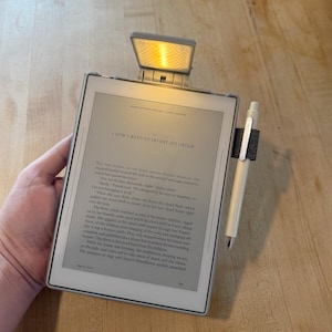 Può includere: Un e-reader grigio con uno schermo rettangolare che mostra il testo intitolato I DON'T NEED TO INVENT ICE CREAM. Una piccola luce regolabile e illuminata è fissata al bordo superiore e una penna color crema è tenuta in un passante di tessuto scuro.