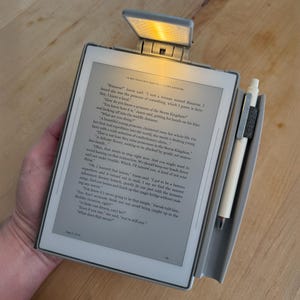 May include: A grey e-reader with a digital screen displaying text, featuring a clip-on light at the top emitting a warm yellow glow and a white stylus pen held in a side loop. The screen shows a narrative passage titled A LIPS ADVENTURE.